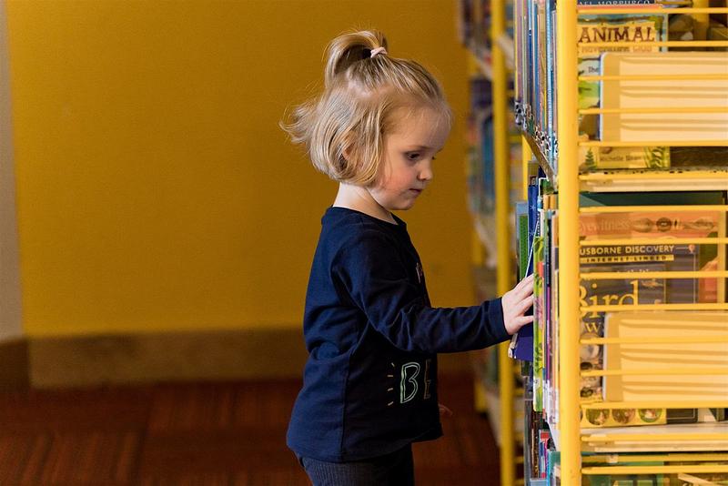 Little girl taking a picture book from yellow, metal bookshelves